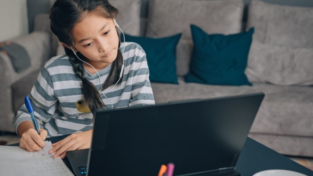 Primary school girl taking notes during a live online tuition class on her laptop at home